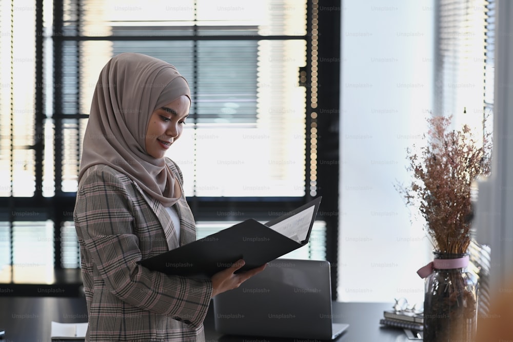 A happy islamic girl in hijab is inspecting and approving documents in folder and standing beside windowpanes in office.
