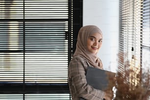 Portrait of muslim businesswoman in hijab is holding document and smiling to camera while standing by the window.