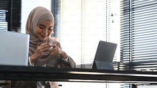 Eine muslimische Geschäftsfrau im Hijab sitzt an ihrem Schreibtisch im Büro und trinkt Kaffee oder Tee.