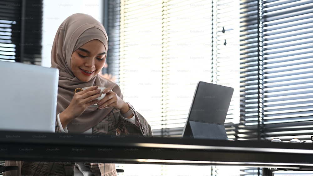 Una donna d'affari musulmana in hijab seduta alla sua scrivania in ufficio e beve caffè o tè.