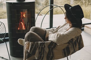Stylish woman in knitted sweater and hat warming up at modern black fireplace with view on mountains. Cozy warm moments at cold season. Hipster female relaxing in comfortable home