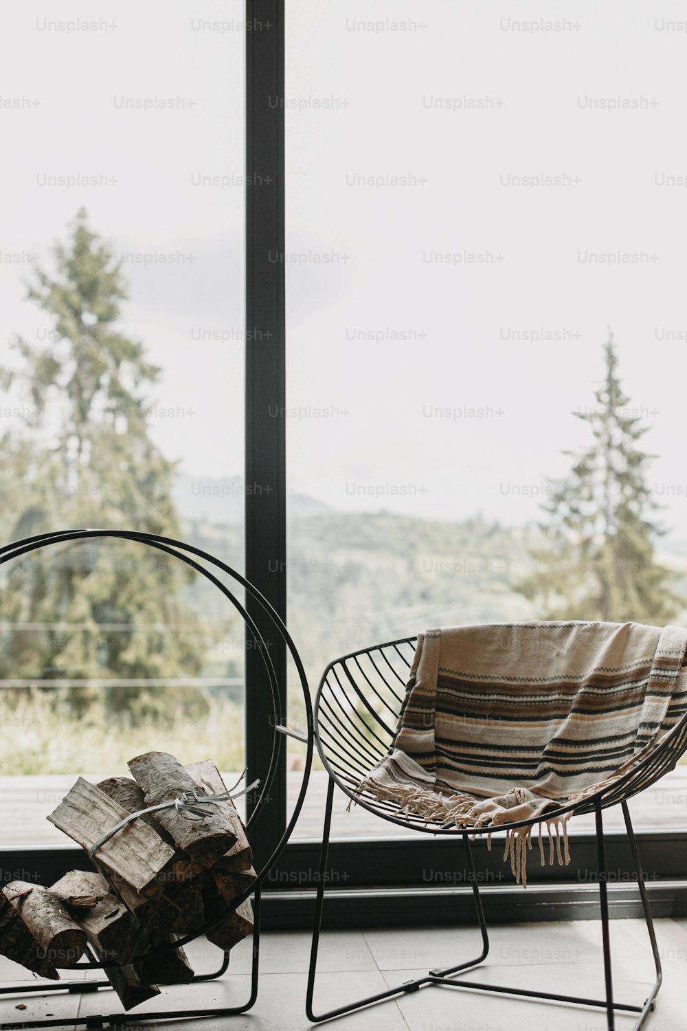 Moderner schwarzer Stuhl mit kuscheliger Decke und Brennholz auf Metallständer vor dem Hintergrund großer Fenster mit Blick auf die Berge. Gemütliche warme und ruhige Momente in der Kabine in der kalten Jahreszeit.