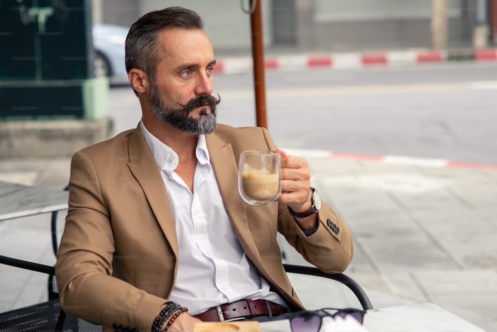 Porträt eines kaukasischen Bartmannes, der mit einer Tasse Kaffee auf dem Tisch vor dem Café sitzt.