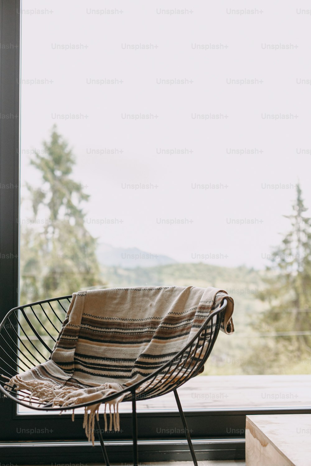 山々を望む大きな窓の背景には、居心地の良い毛布と金属の上の薪が置かれたモダンな黒い椅子が置かれています。寒い季節のキャビンでの居心地の良い暖かく穏やかなひととき。