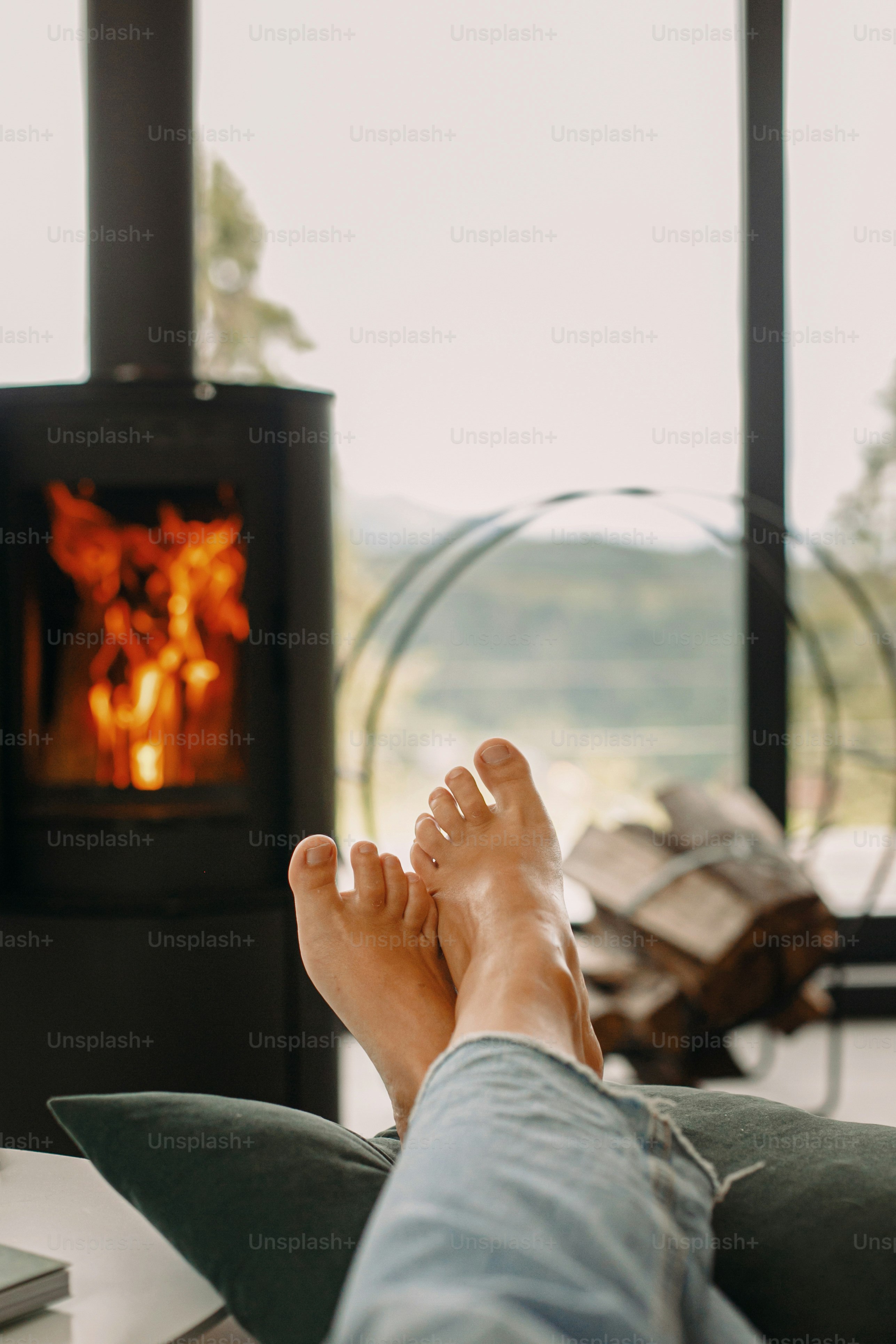 Kaki di latar belakang perapian hitam modern dan jendela besar dengan pemandangan pegunungan. Wanita bertelanjang kaki bersantai di rumah yang nyaman, momen hangat yang nyaman