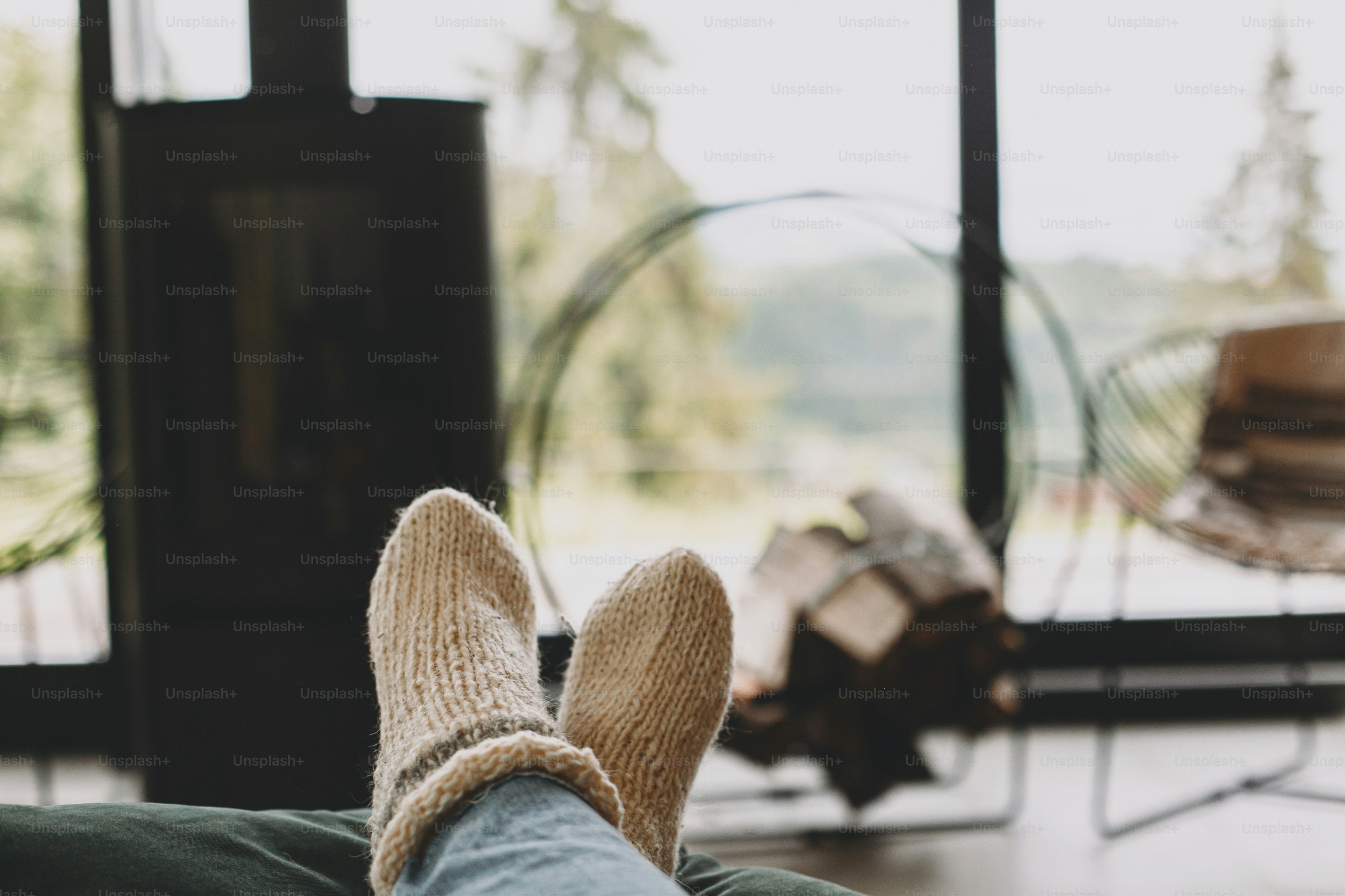 Feet in warm knitted woolen socks on background of modern black fireplace and big windows with view on mountains. Woman relaxing in comfortable home, cozy warm moments