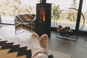 モダンな黒い暖炉と山々の景色を望む大きな窓を背景に、暖かいニットのウールの靴下の足。快適な家でリラックスする女性、居心地の良い暖かい瞬間