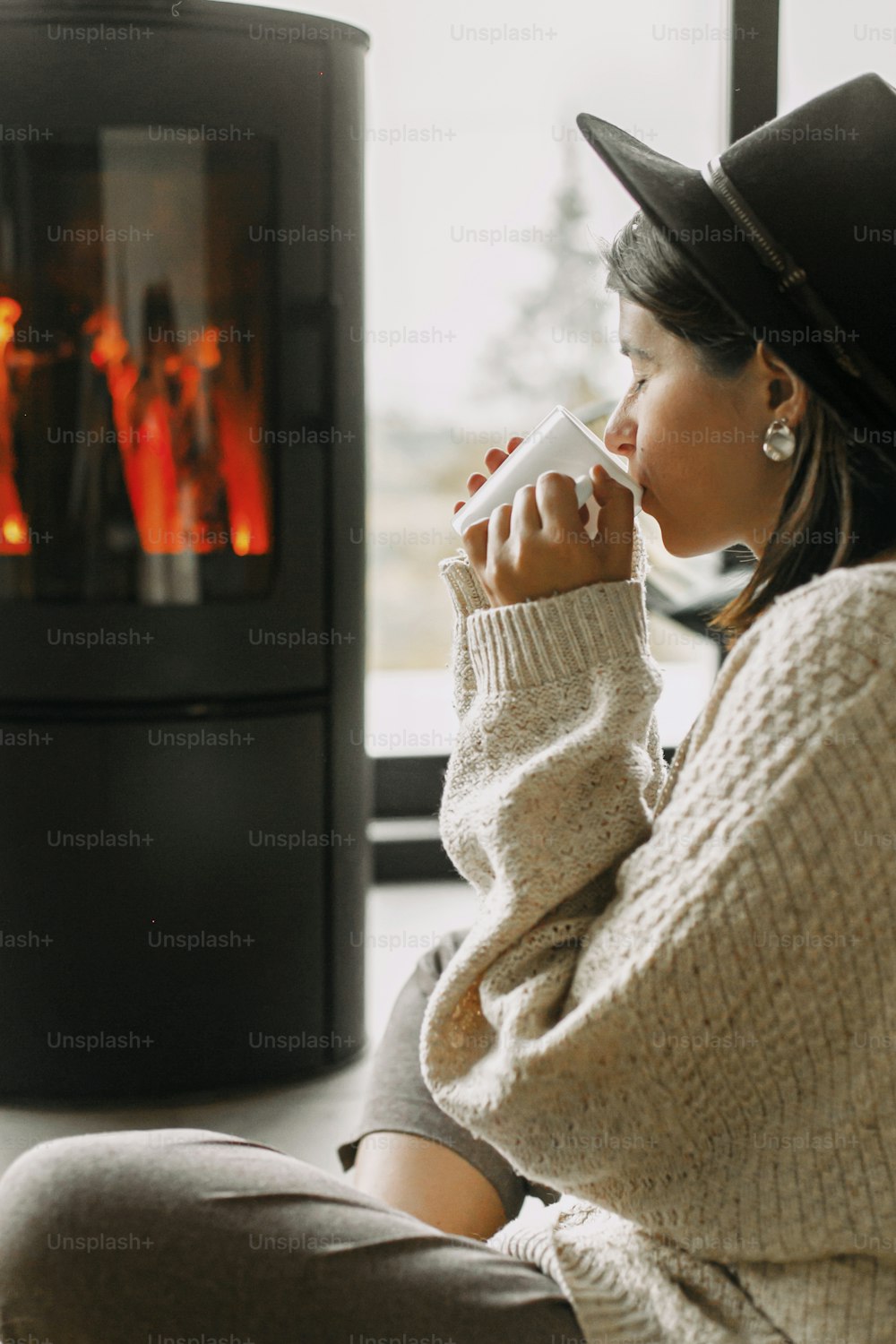Stylish woman in knitted sweater drinking warm cup of tea and relaxing at modern black fireplace with view on mountains. Cozy warm moments at cold season. Young female enjoying drink