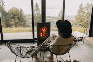 Stilvolle Frau in Strickpullover und Mütze mit warmer Tasse Tee am modernen schwarzen Kamin mit Blick auf die Berge. Gemütliche warme Momente in der kalten Jahreszeit. Junge Hipster-Frau entspannt sich in gemütlichem Zuhause