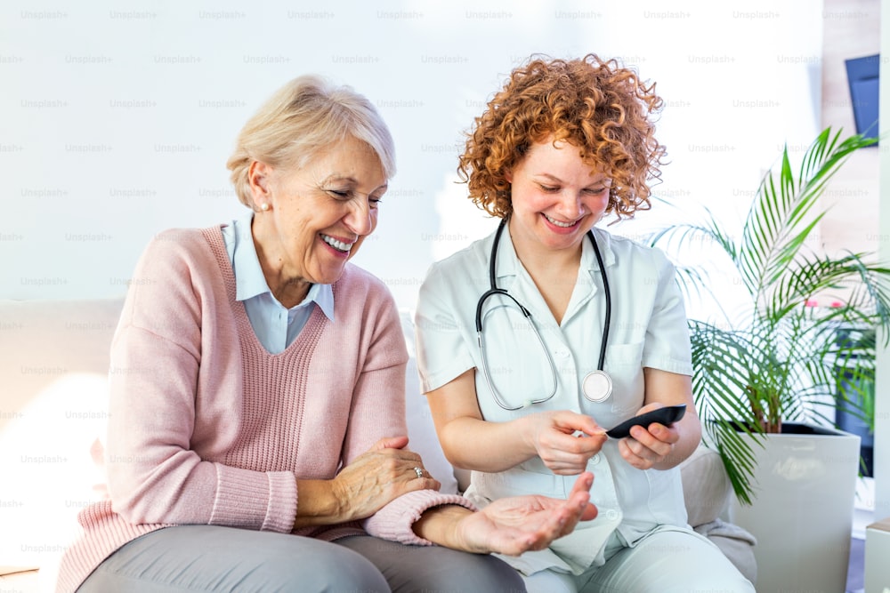 Femme âgée heureuse de se faire mesurer sa glycémie dans une maison de retraite par son aide-soignante. Infirmière heureuse mesurant la glycémie d’une femme âgée dans le salon - concept de diabète et de glicémie