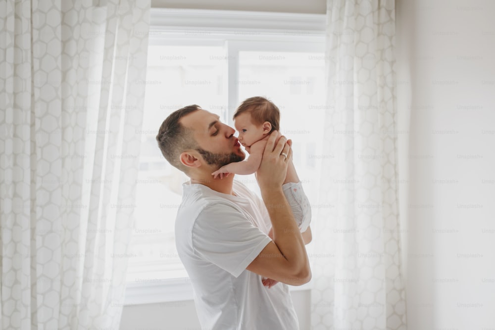 Happy Caucasian father kissing newborn baby. Male man parent holding rocking child daughter son on his hands. Authentic lifestyle candid moment. Young bearded dad family life concept.
