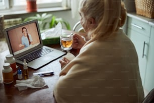 Senior lady holding cup of tea and having online consultation with female therapist