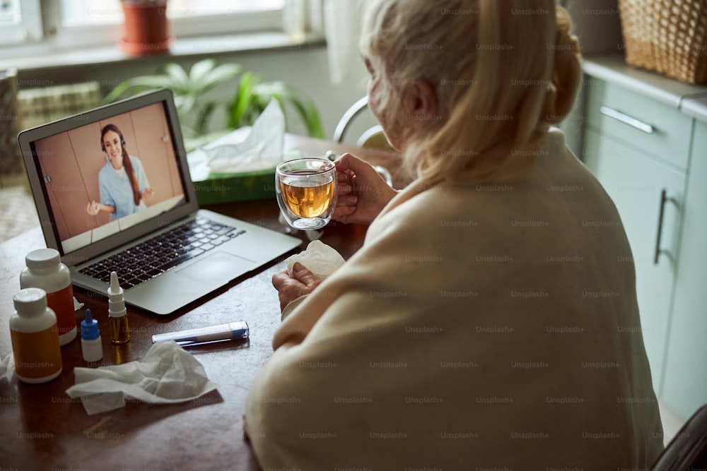 Senior lady holding cup of tea and having online consultation with female therapist