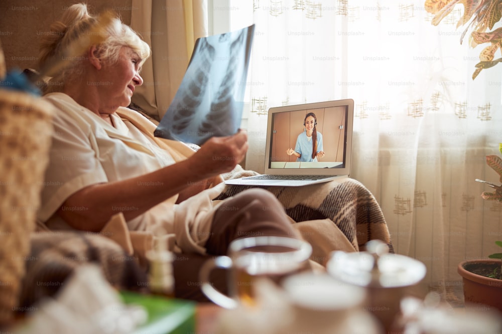 Elderly lady sitting in armchair and holding chest x-ray while talking with female physician through video call