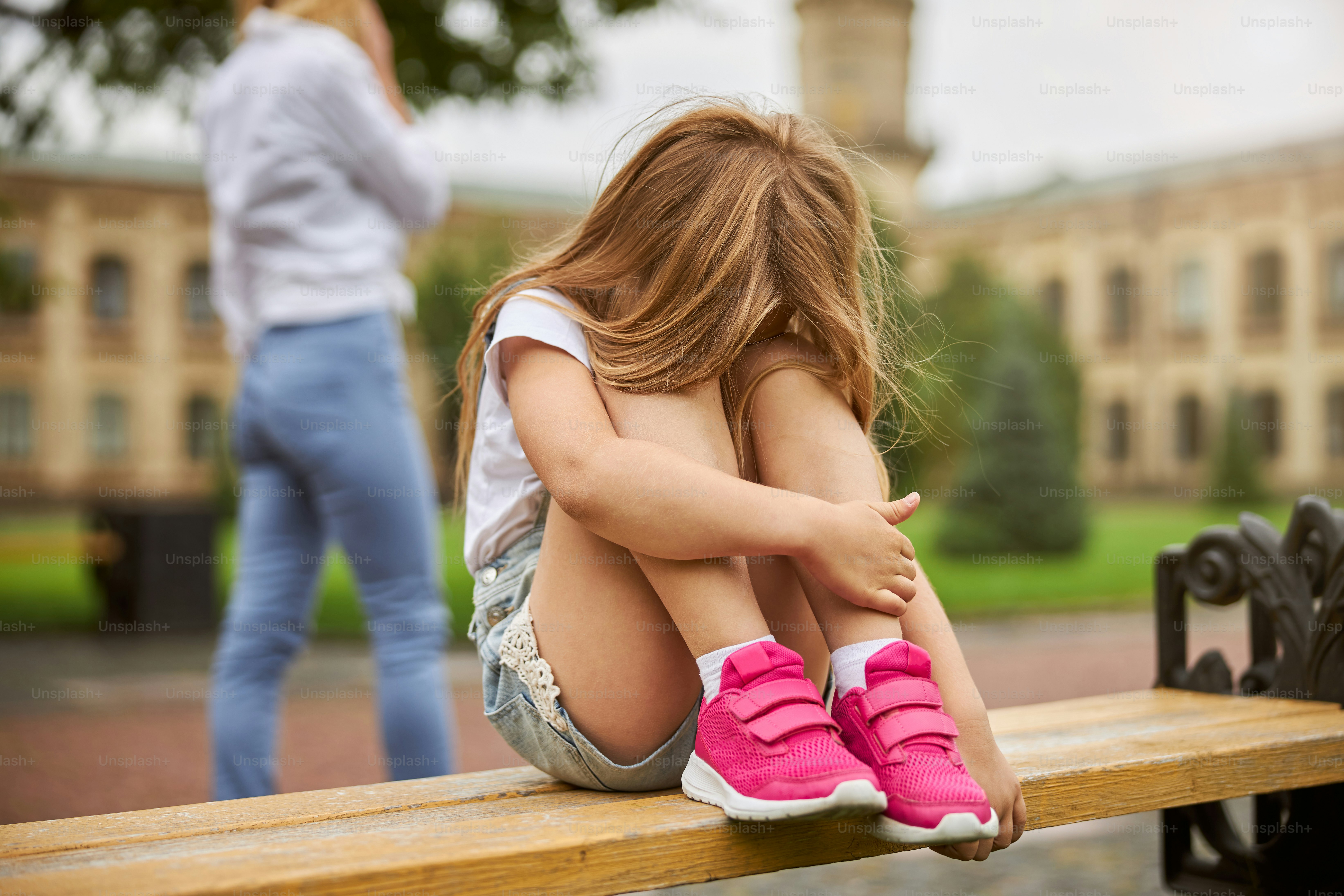 Young girl sitting in a playground in the day with her face in her lap because she is sad