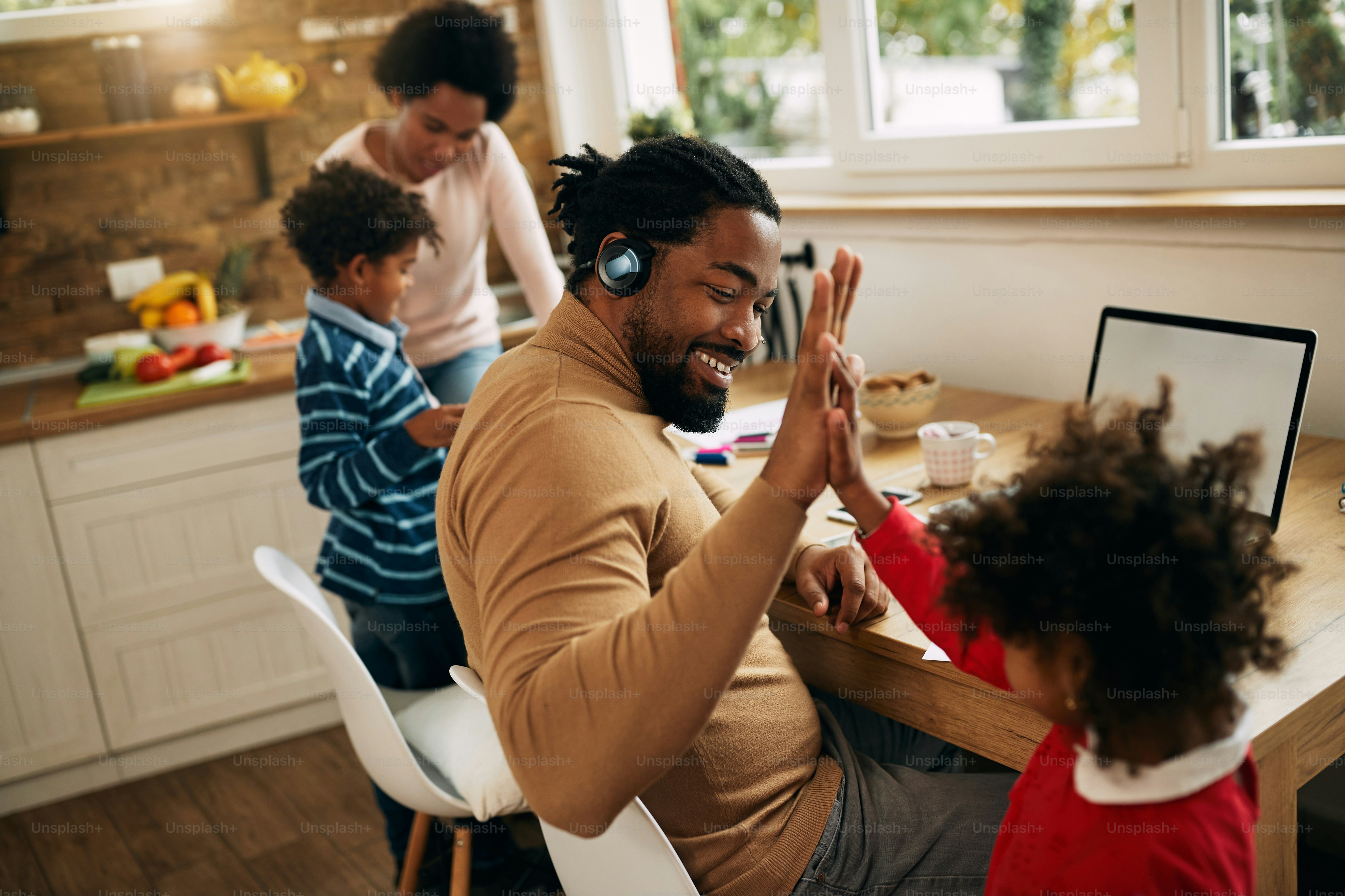 Homem afro-americano feliz dando high-five para sua filha pequena enquanto trabalhava em laptop em casa.