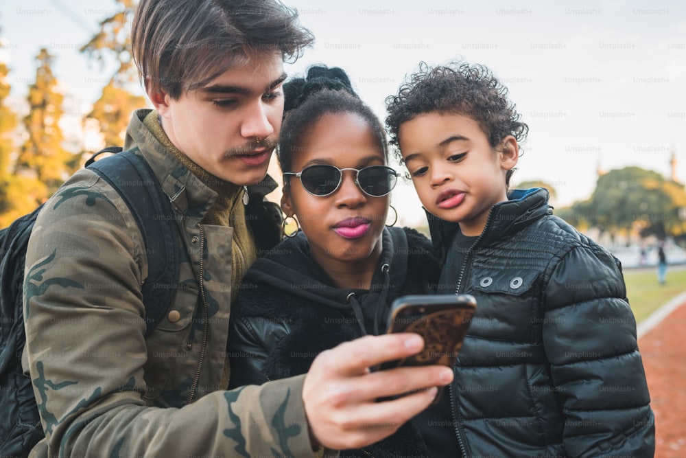 Retrato de una encantadora familia étnica de raza mixta divirtiéndose, relajándose y usando el teléfono móvil en el parque al aire libre.