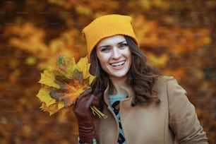 Hello november. smiling stylish middle aged woman in beige coat and orange hat with autumn yellow leaves outside in the city park in autumn.