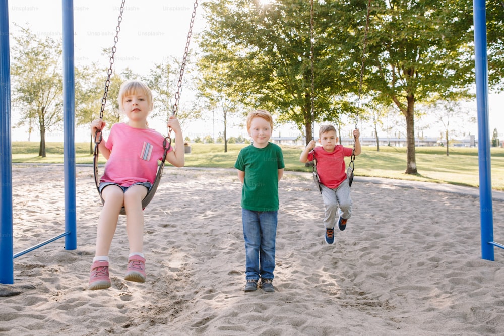 Happy smiling little preschool girl and boys friends swinging on swings at playground outside on summer day. Happy childhood lifestyle concept. Seasonal outdoor activity for kids.