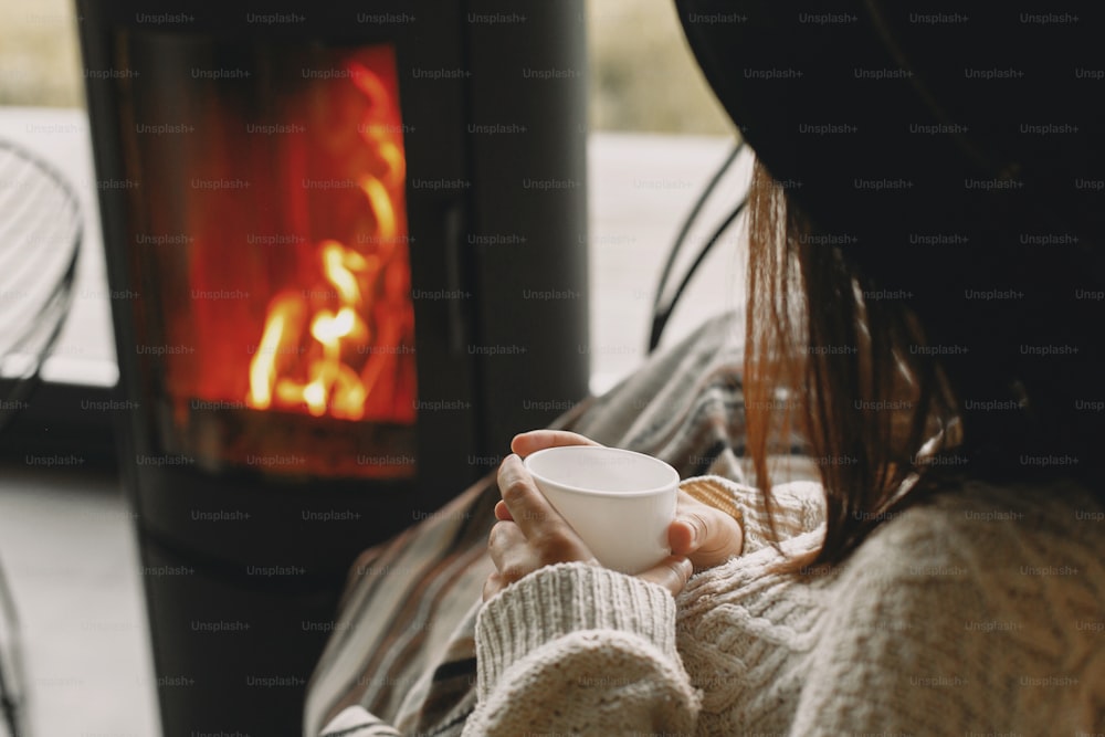 Stylish woman holding warm cup of tea and relaxing at modern black fireplace with view on mountains. Cozy warm moments at cold season. Young female in knitted sweater and hat resting
