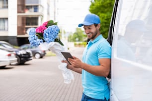 Happy courier with a tablet PC during flowers delivery is waiting for a client