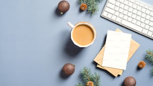 Espaço de trabalho feminino com xícara de café, teclado, cartão de papel em branco, pinhas e galhos, bolas sobre fundo azul pastel. Natal, conceito de férias de inverno. Aconchegante, hygge, estilo nórdico.