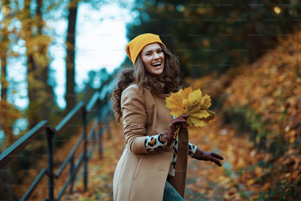 Hello november. happy trendy woman in brown coat and yellow hat with autumn yellow leaves enjoying promenade outside in the city park in autumn.
