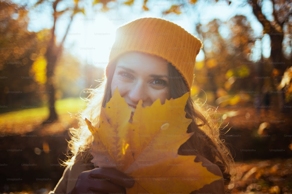 Hola octubre. Mujer moderna feliz de 40 años en abrigo beige y sombrero naranja con hojas amarillas otoñales afuera en el parque de la ciudad en otoño.