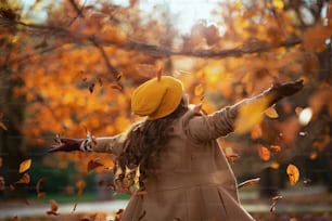Ciao autunno. Visto da dietro, donna di mezza età in cappotto beige e cappello arancione che si rallegra all'aperto nel parco cittadino in autunno.