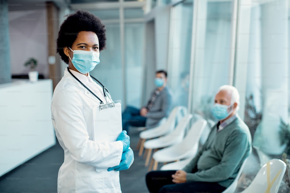 Portrait of black female doctor wearing protective face mask and looking at camera while standing in a waiting room at the hospital. There are people in the background.