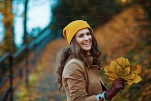 안녕하세요 가을입니다. 갈색 코트와 노란 모자를 쓴 행복한 세련된 여자는 가을에 도시 공원에 밖에 가을 노란 잎을 가지고 있다.
