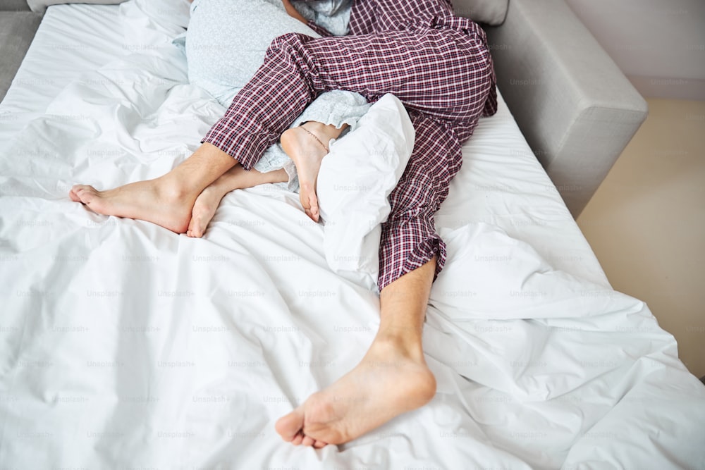 Close up of barefoot man in pajamas hugging girlfriend while resting on white sheets