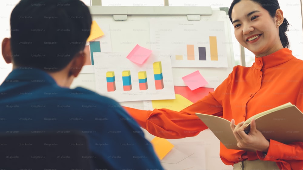 Junge Frau erklärt Geschäftsdaten auf Whiteboard in zwanglosem Büroraum. Die selbstbewusste asiatische Geschäftsfrau berichtet über den Informationsfortschritt eines Geschäftsprojekts an Partner, um die Marktstrategie festzulegen.