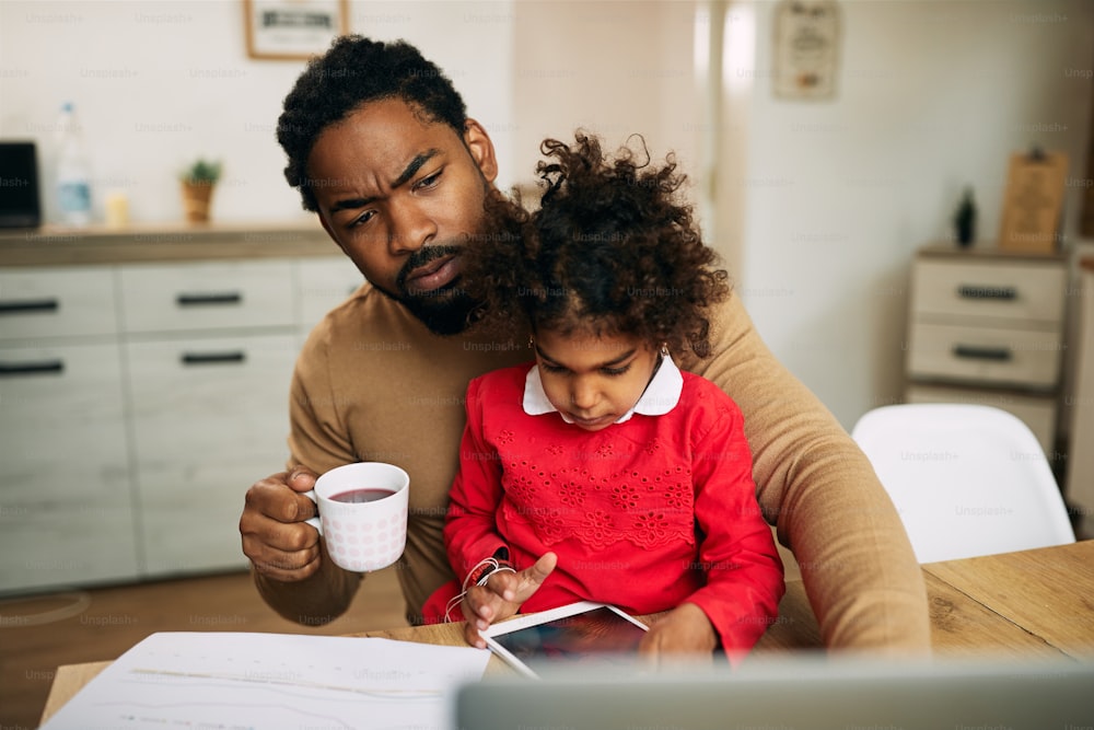 Black working father drinking tea and using laptop while his daughter is sitting in his lap and using digital tablet.