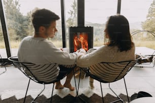 Stylish couple in sweaters with warm cups of tea relaxing at modern black fireplace with view on mountains. Cozy warm family moments at cold season. Young family on vacation