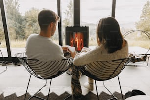 Stylish couple in sweaters with warm cups of tea relaxing at modern black fireplace with view on mountains. Cozy warm family moments at cold season. Young family on vacation