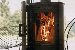 Modern black fireplace with burning fire and firewood on metal stand on background of big window. Cozy warm and calm moments at cold season, heater in cabin