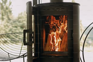 Moderner schwarzer Kamin mit brennendem Feuer und Brennholz auf Metallständer auf dem Hintergrund des großen Fensters. Gemütliche warme und ruhige Momente in der kalten Jahreszeit, Heizung in der Kabine