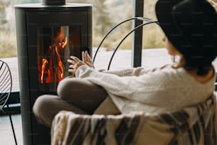 ニットのセーターと帽子をかぶったスタイリッシュな女性が、山々を眺めながらモダンな黒い暖炉で暖めています。寒い季節に心地よい温かいひとときを。快適な家でリラックスするヒップスターの女性