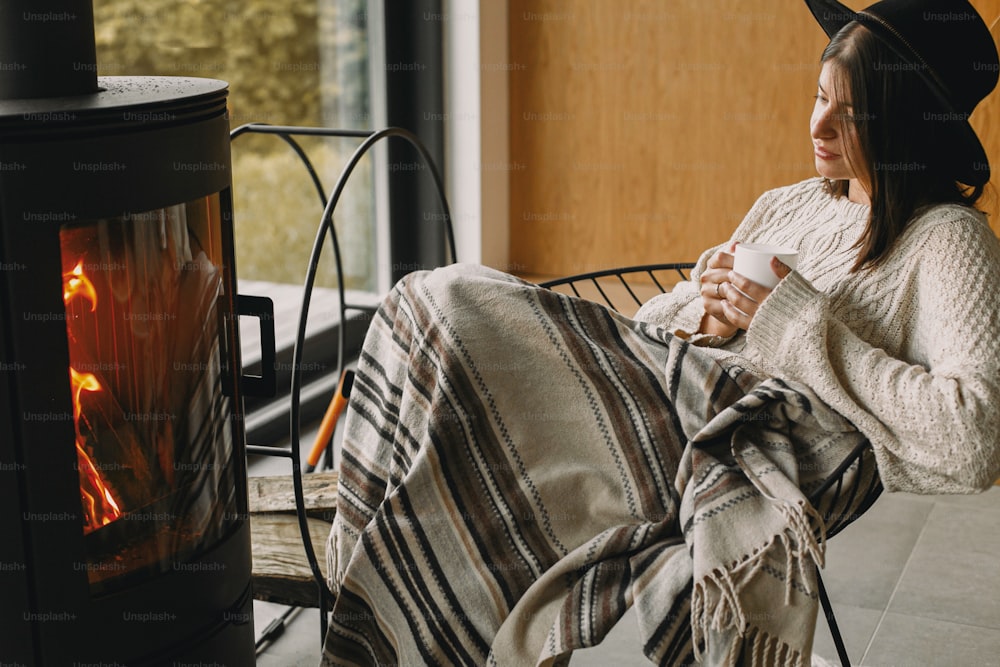 Stylish woman in knitted sweater and hat holding warm cup of tea at modern black fireplace with view on mountains. Cozy warm moments at cold season. Young hipster female relaxing in comfy home