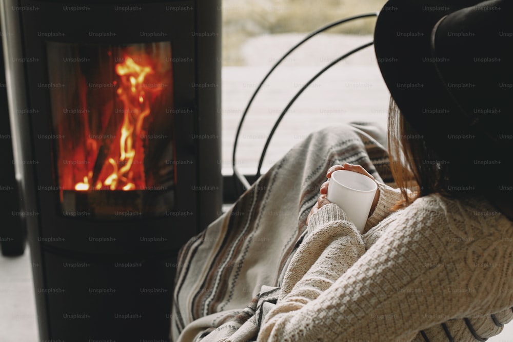 Stylish woman holding warm cup of tea and relaxing at modern black fireplace with view on mountains. Cozy warm moments at cold season. Young female in knitted sweater and hat resting