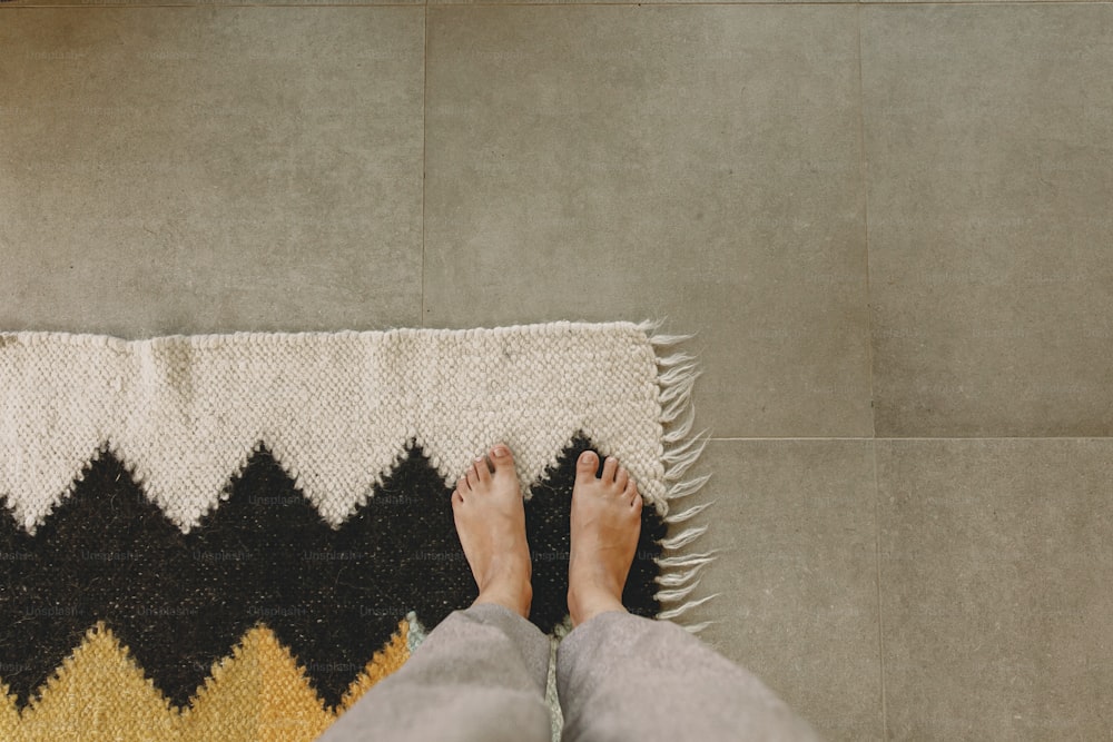 Woman barefoot standing on modern rug in comfortable stylish home, top view. Feet on cozy woolen rug on background of concrete floor. Cozy warm moments