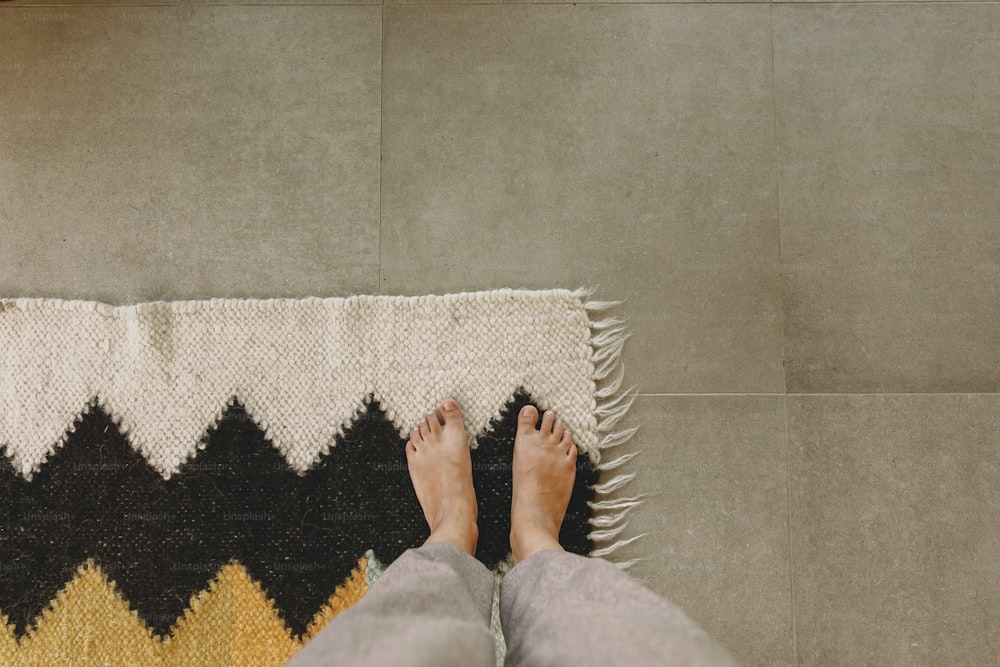 Mujer descalza de pie sobre alfombra moderna en una casa cómoda y elegante, vista superior. Pies sobre acogedora alfombra de lana sobre fondo de piso de hormigón. Momentos cálidos y acogedores