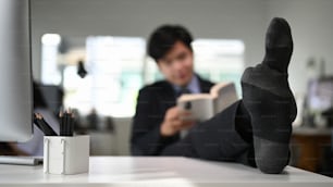 A businessman in black suit is put his feet on the desk and reading ao book in his office room.
