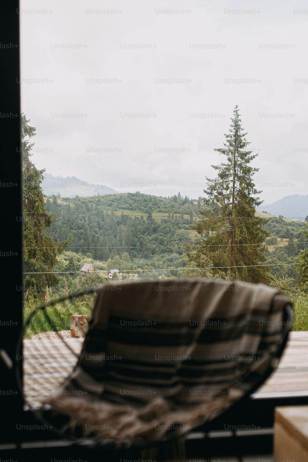 Beautiful view on mountains hills and trees through window cabin. Blurred modern metal chair with cozy blanket on background of big window and trees in focus