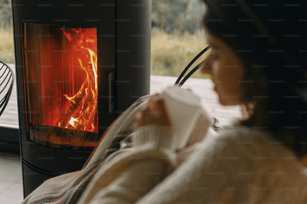 モダンな黒い暖炉で燃える火と、ニットセーターを着た女性が温かいお茶を飲んでリラックスしているぼやけたイメージ。寒い季節に心地よい温かいひとときを