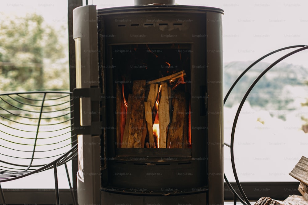 Camino nero moderno con fuoco ardente e legna da ardere su supporto metallico sullo sfondo della grande finestra. Momenti accoglienti caldi e tranquilli nella stagione fredda, riscaldamento in cabina