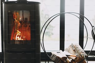 Modern black fireplace with burning fire and firewood on metal stand on background of big windows with view on mountains. Cozy warm  and calm moments at cold season.