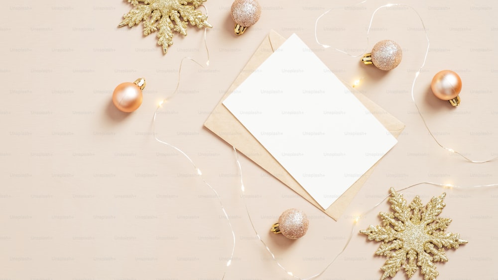 파스텔 베이지색 배경에 빈 흰색 카드 모형, 황금 눈송이, 공이 있는 종이 봉투 편지. 우아한 크리스마스 플랫 레이 구성, 상위 뷰.