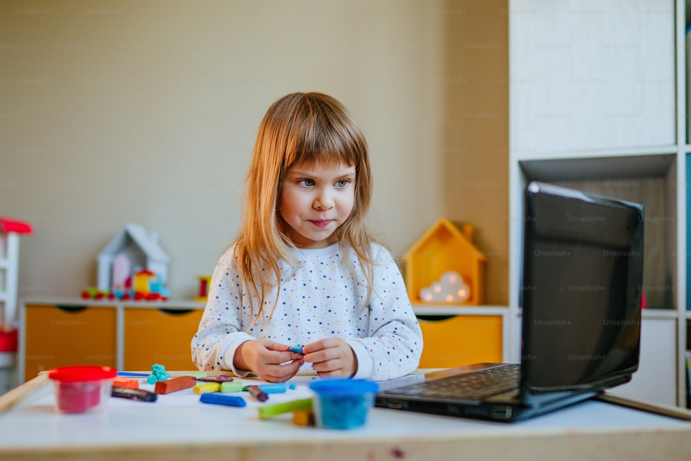 Kleines Mädchen formt bunten Ton und schaut sich Online-Lernstunde auf dem Laptop drinnen an. Fernunterrichtskonzept.